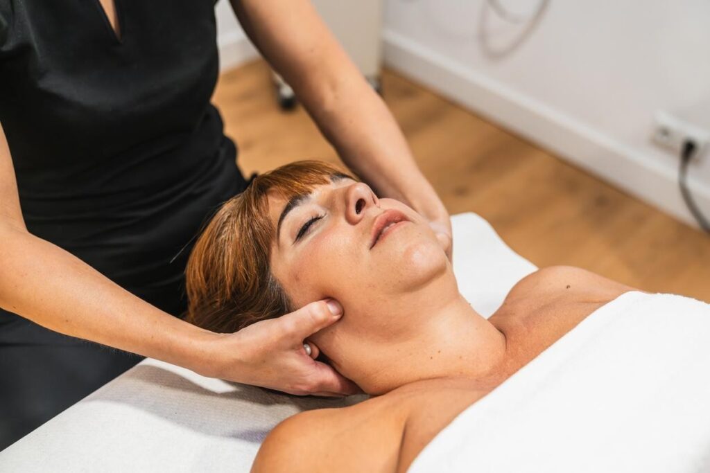 TMJ Massage Downtown Brooklyn: A woman with closed eyes is receiving a facial or neck massage from a therapist in a clean, brightly lit treatment room.