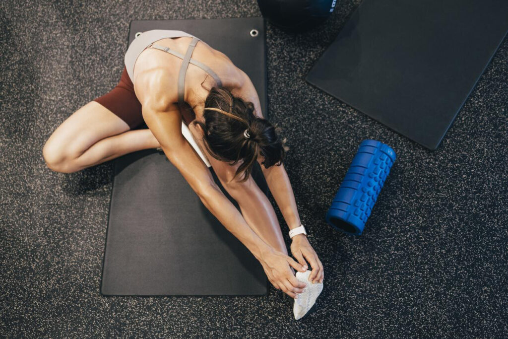 Sports Massage for Injury Prevention: A woman in athletic wear sits on a gym mat, reaching forward to stretch her leg next to a blue foam roller.