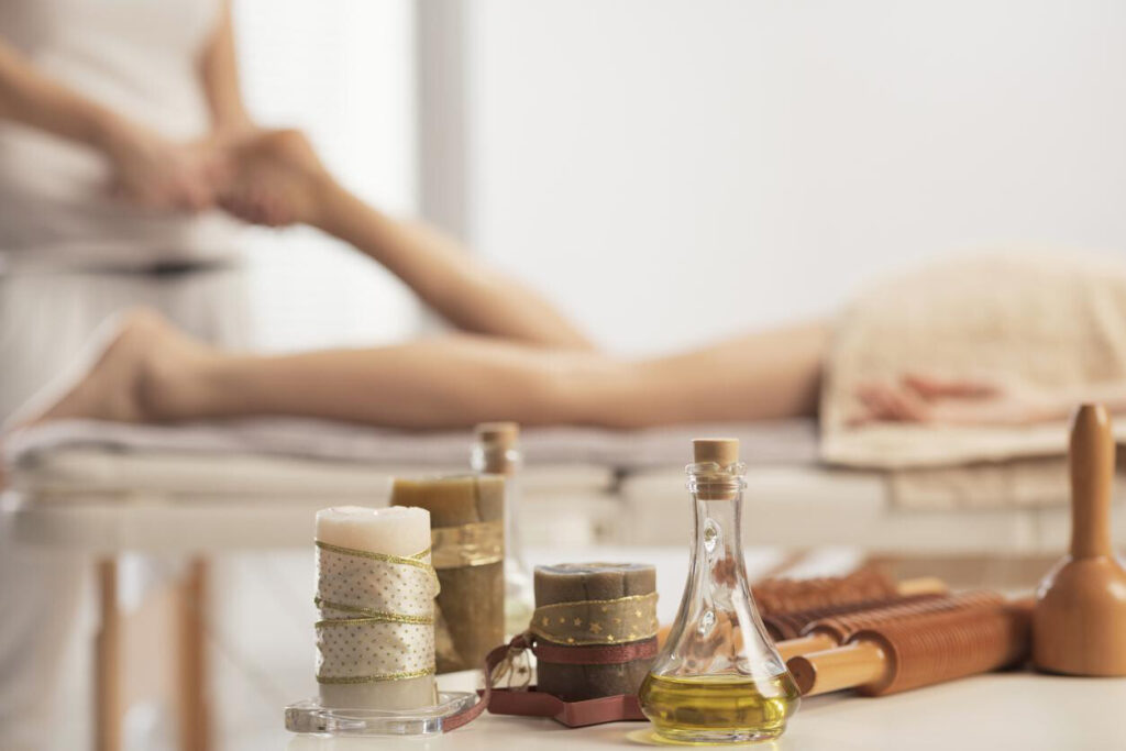 Sports Massage for Injury Prevention: In a shallow focus shot, a glass bottle of oil, decorative candles, and wooden massage rollers sit on a table while a massage therapist works in the blurred background.