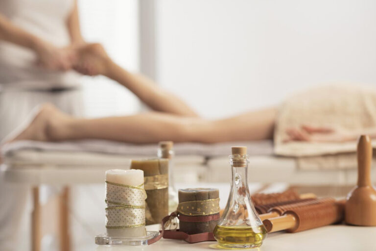 Sports Massage for Injury Prevention: In a shallow focus shot, a glass bottle of oil, decorative candles, and wooden massage rollers sit on a table while a massage therapist works in the blurred background.